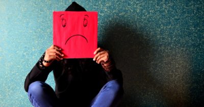 A mid height shot of a person sat against a blue wall, holding a red piece of paper in front of them with a sad face drawn on it