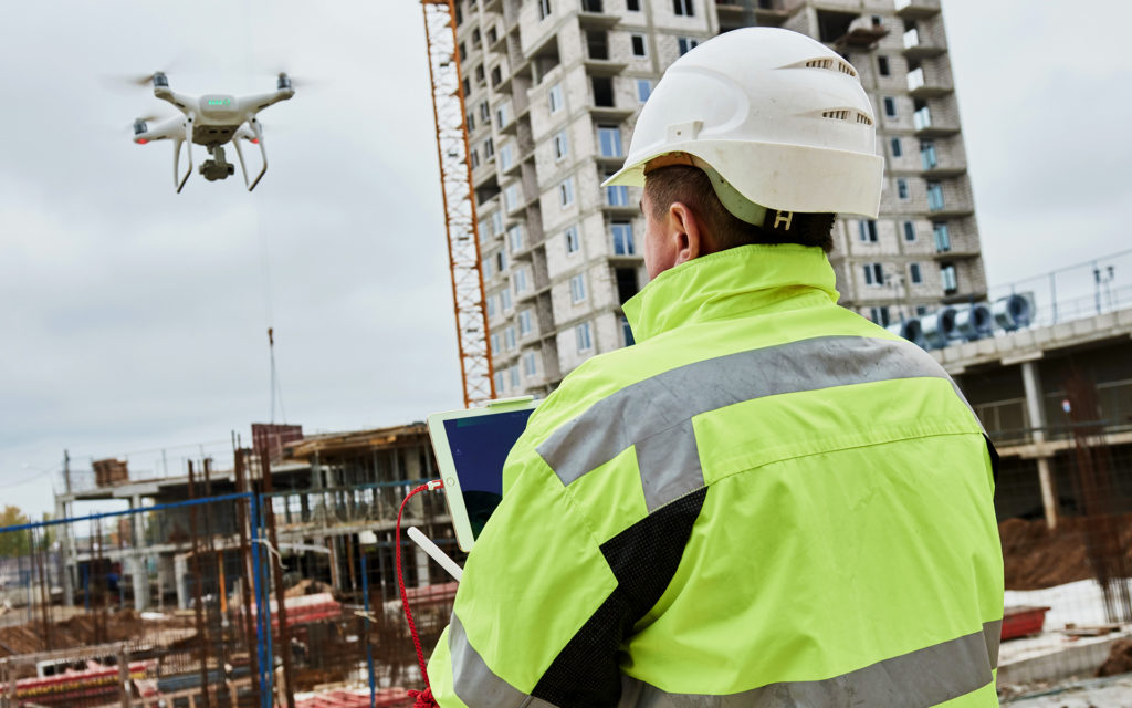 An over the shoulder shot of a male construction worker wearing a white hard hat, controlling a drone with a construction site in the background