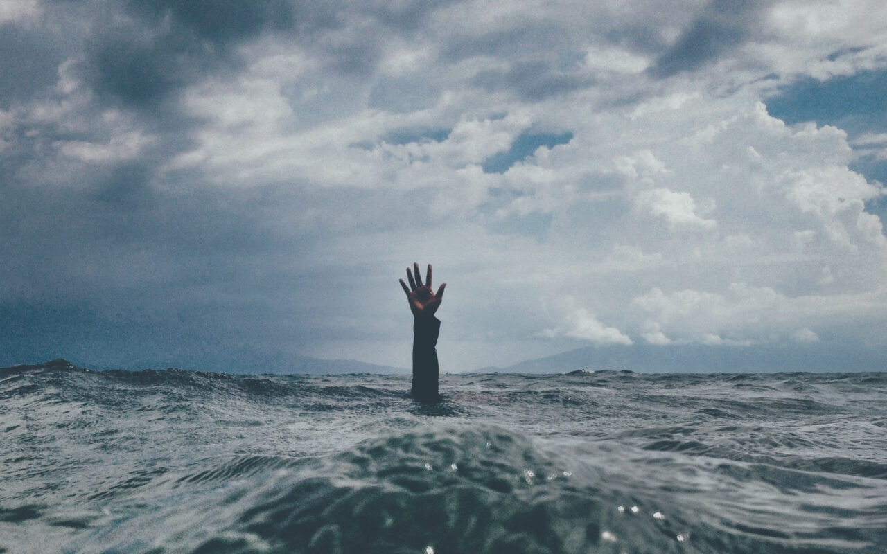 A low angle shot of a hand reaching out from underneath an ocean wave, with grey clouds in the background