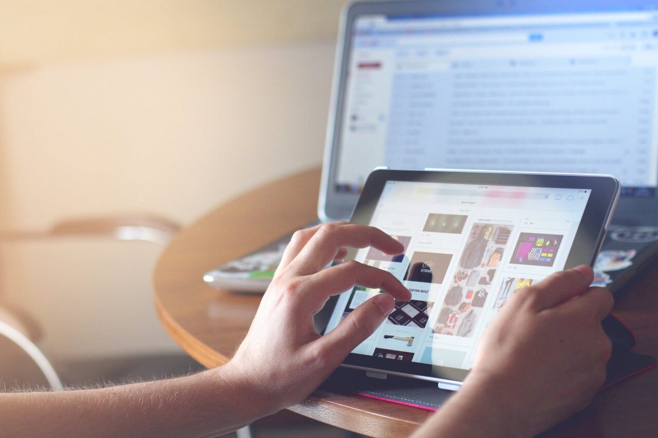 An image of hands using an iPad with multiple images on it, in front of an out of focus laptop.