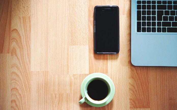 A birds-eye-view photo of a mobile phone next to a coffee cup and a laptop on a light brown desk