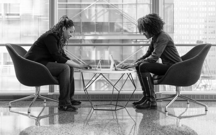 A mid height shot of two women sat on chairs facing one another over a coffee table, both are leaning over electronic tablets