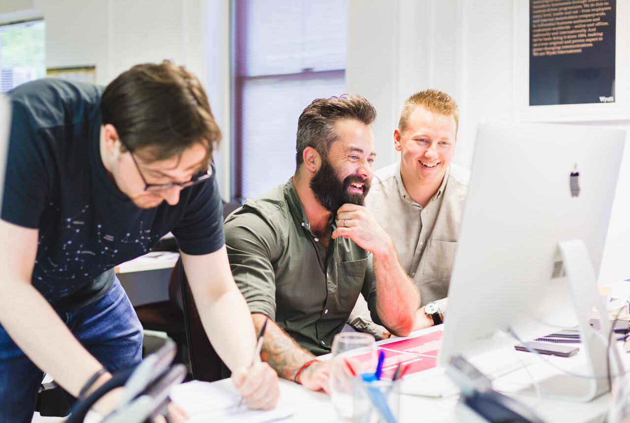 Three male office workers are sat a desk smiling and working in front of a desktop computer screen