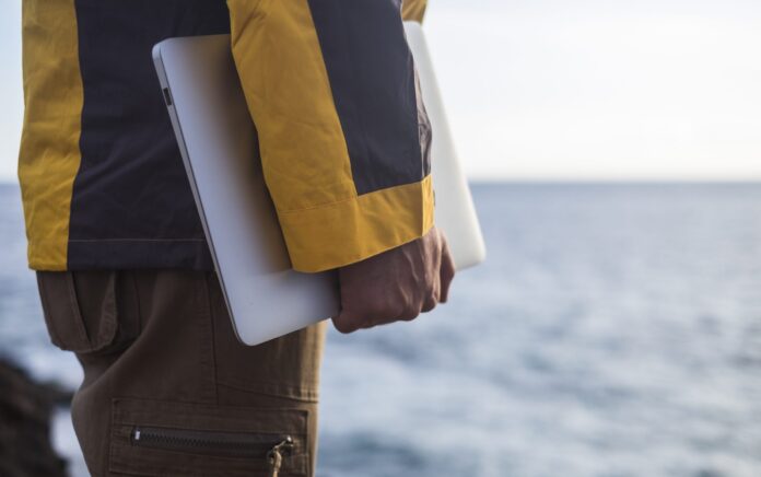 A close up shot of a persons arm holding a laptop, the ocean is blurred out in the background