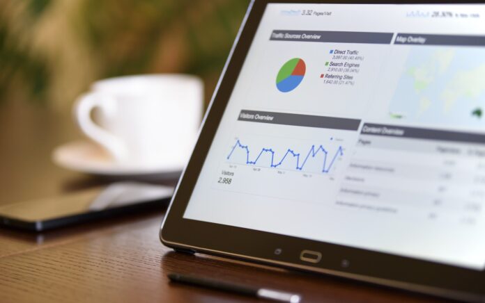 A close up shot of an electronic tablet with various graphs shown on screen sat on top of a wooden desk, a blurred mobile phone and coffee cup are in the background.