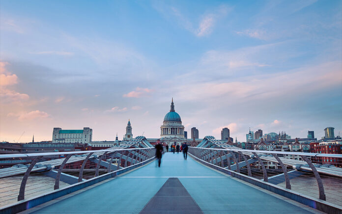 A wide landscape shot of the millennium bridge with blurred pedestrians walking across it, the old bailey is visible in the background
