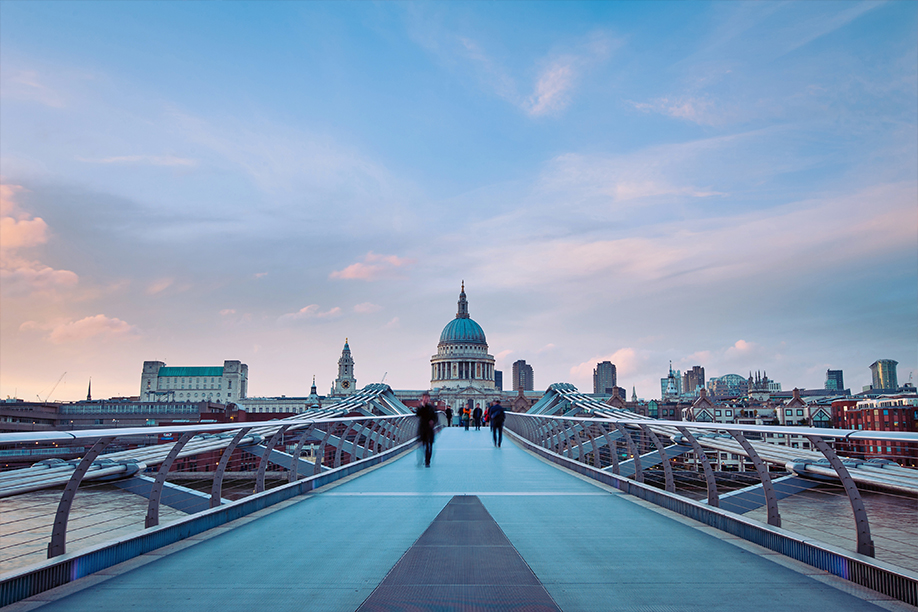 A wide landscape shot of the millennium bridge with blurred pedestrians walking across it, the old bailey is visible in the background