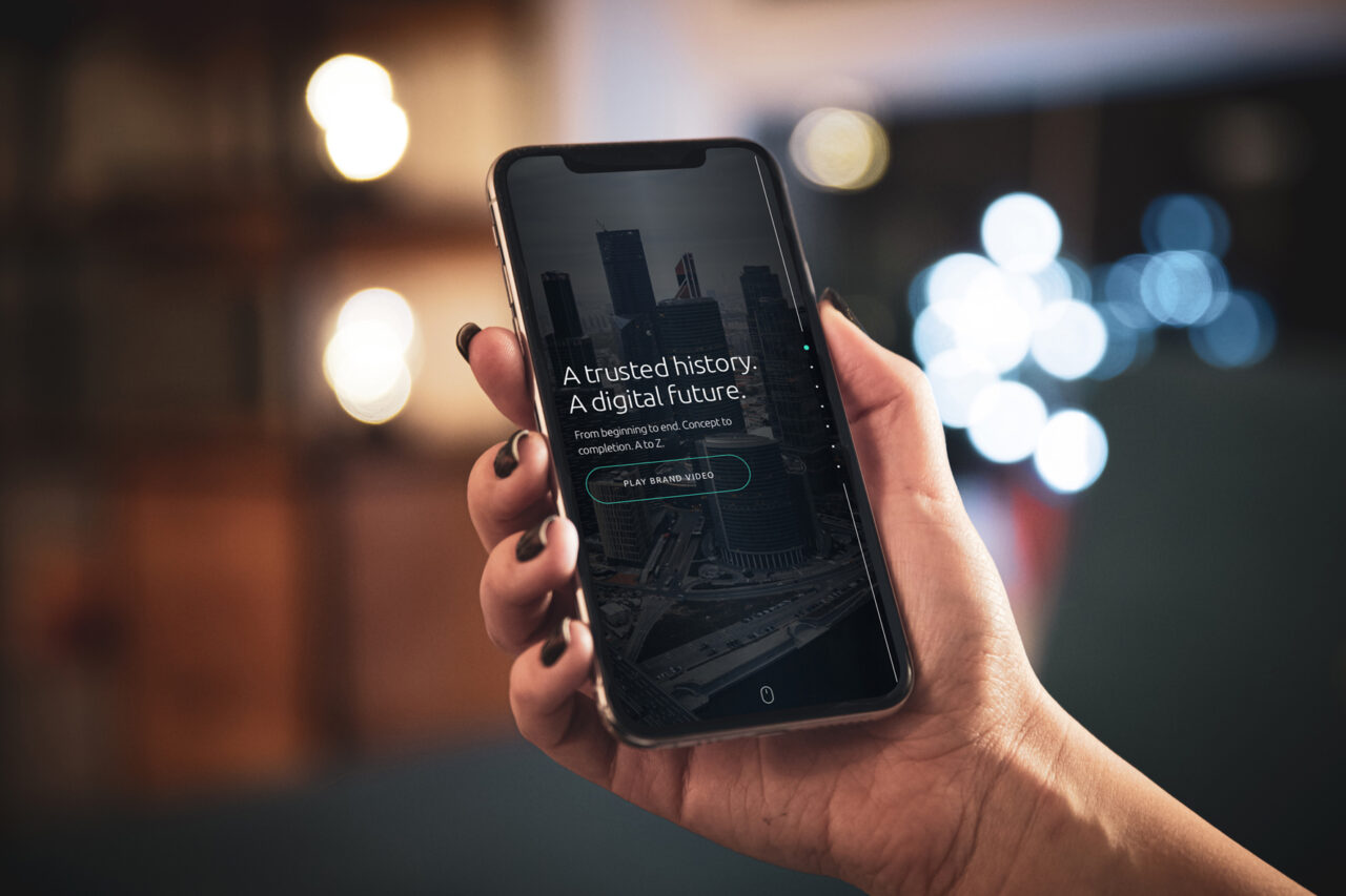 A close up shot of a hand holding a mobile phone with a presentation on screen, blurred lights are shining in the background.