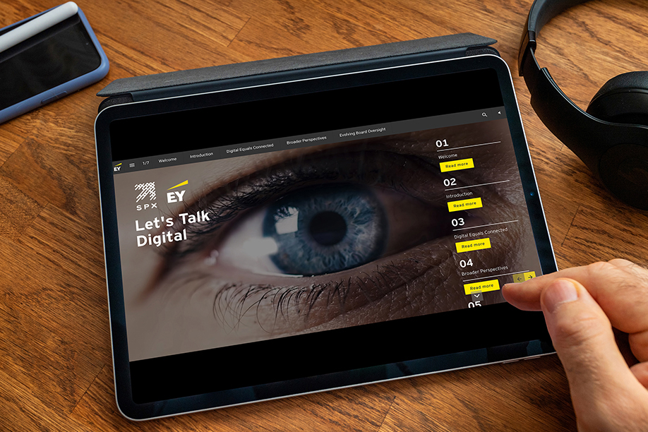 A close up shot of an electronic tablet resting on a brown wooden table, with a mobile phone and headphones next to it. There is a hand pointing at a digital presentation on the tablet screen