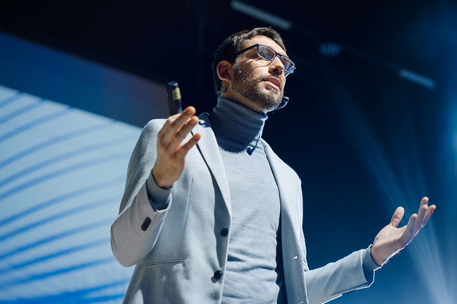 An upwards facing mid range shot of a motivational speaker, wearing a grey suit and lit with blue and silver lighting