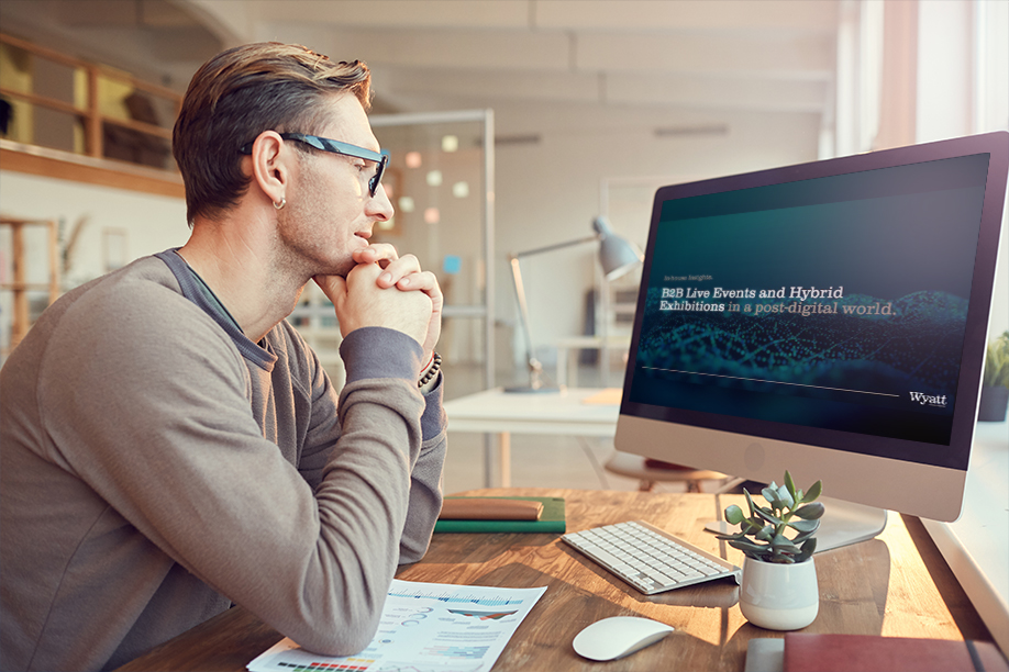 A mid height shot of a male wearing reading glasses, looking at a computer screen situated on top of a desk with paper and a plant on top of it. On the screen is Wyatt International branding.