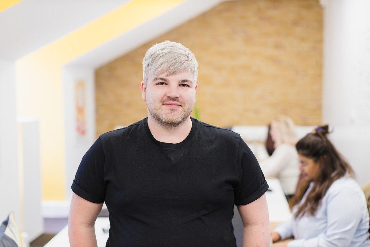 Ben Baker-Hollyhead, Strategic Account Director, stood smiling in an office whilst two colleagues sit a desk in the background