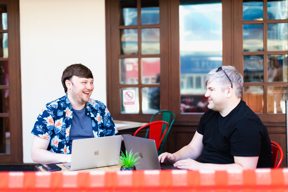 Two males are sat in an outdoor seating area, both are using laptops and smiling at one another