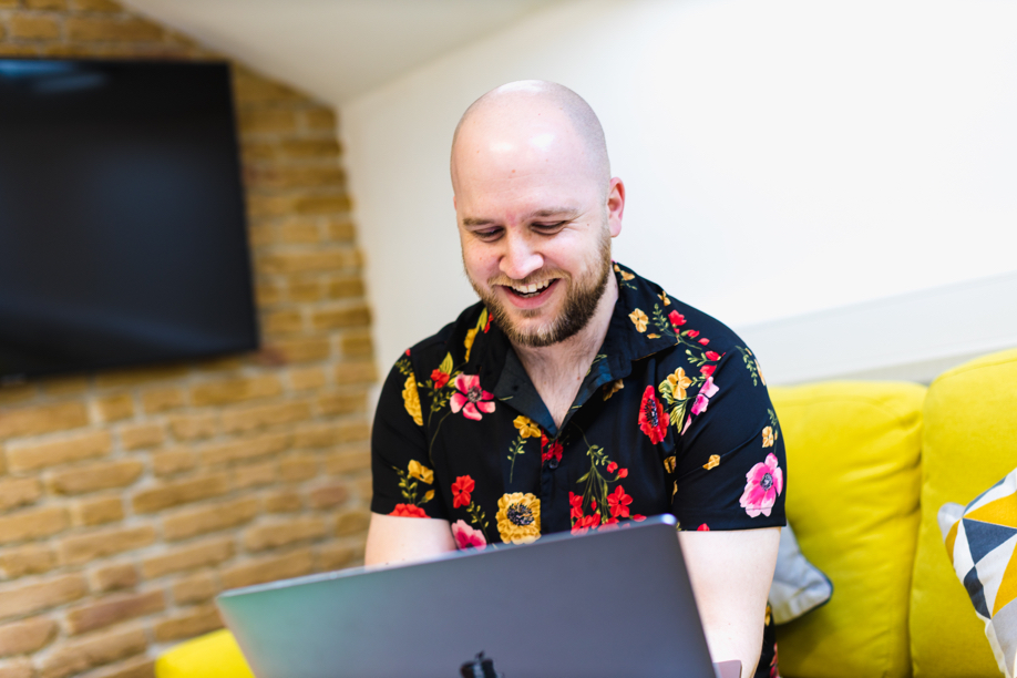 A male is sat on a yellow sofa smiling at a laptop which he is holding