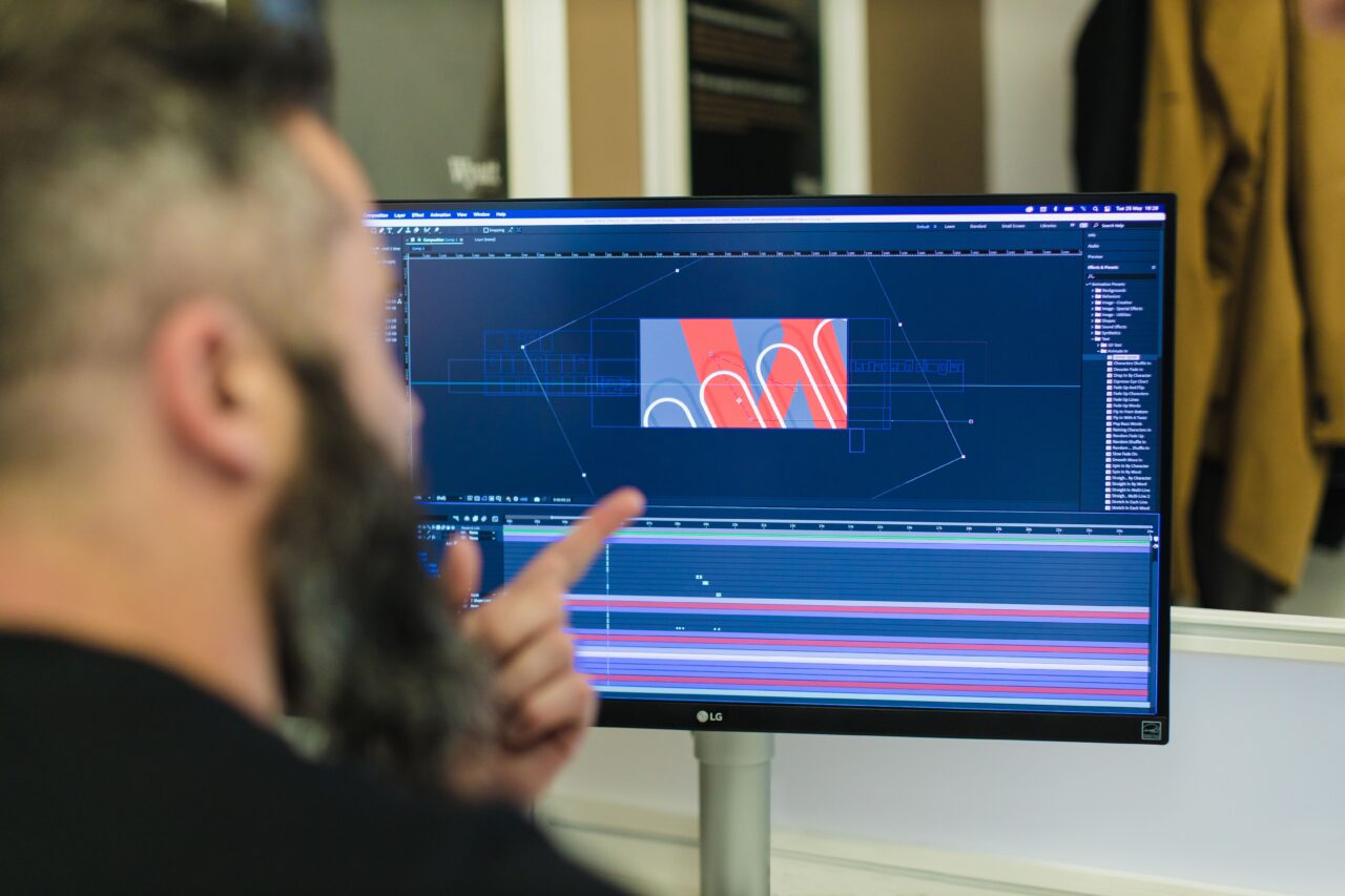 An over the shoulder shot of a male pointing at a computer screen, with graphic design work on screen