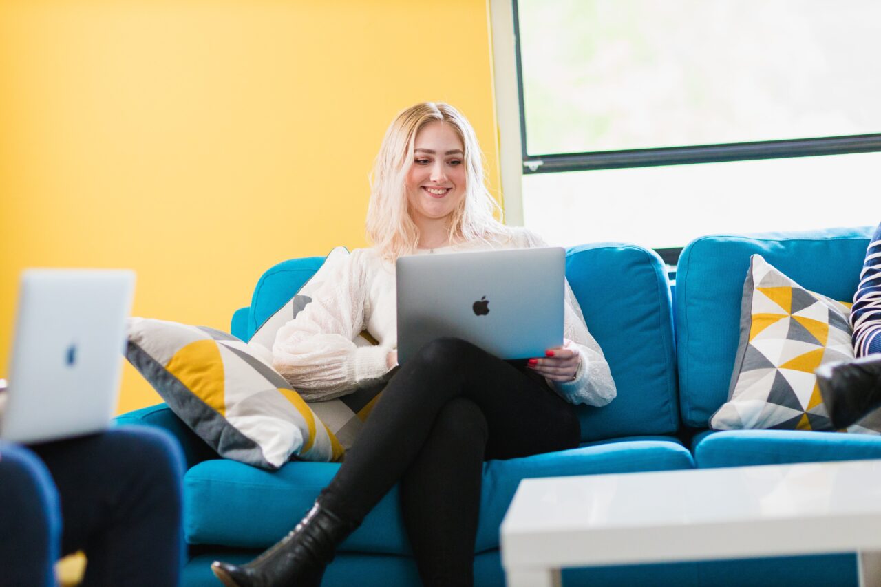 A female office worker is sat on a blue sofa, holding a laptop and smiling