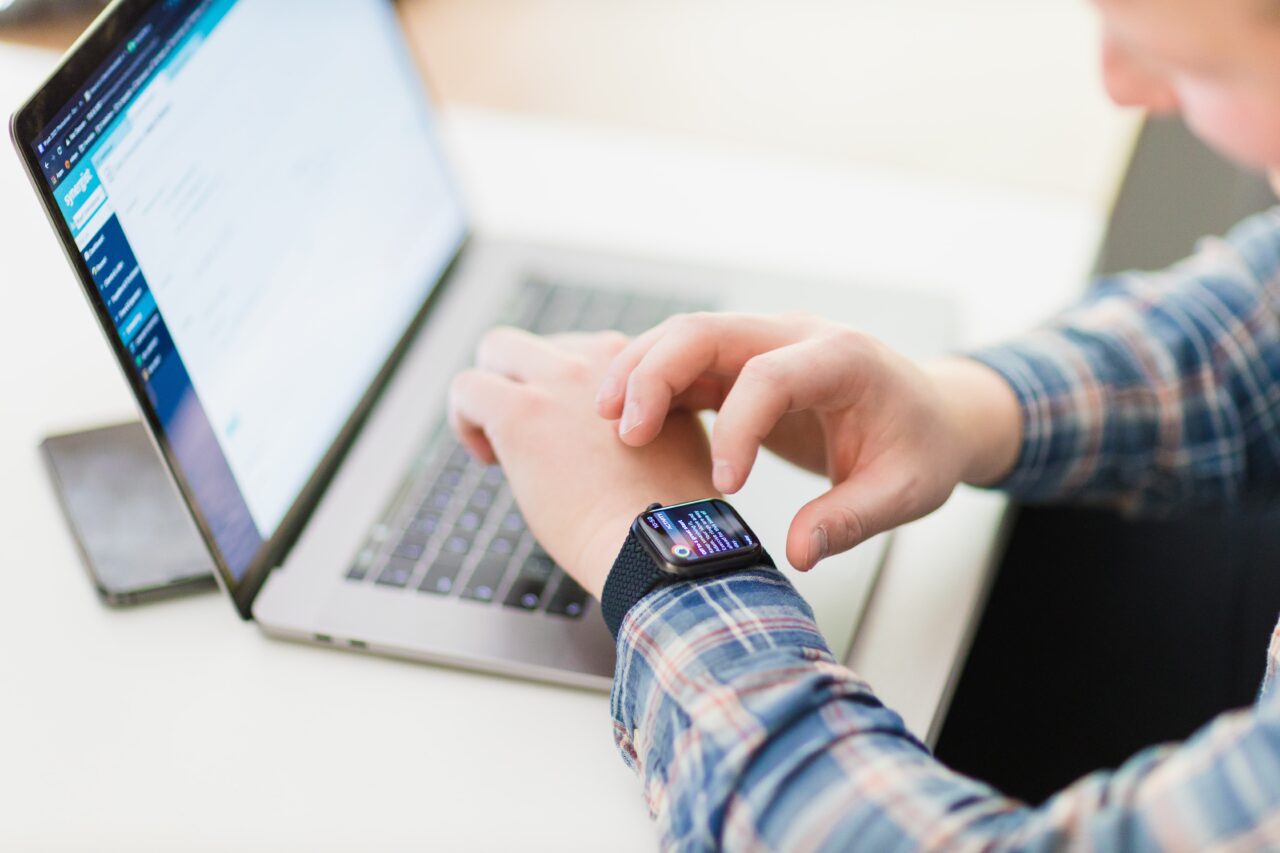 A close up shot taken over the shoulder of a male using an electronic watch, with a laptop sat on the desk in front of him