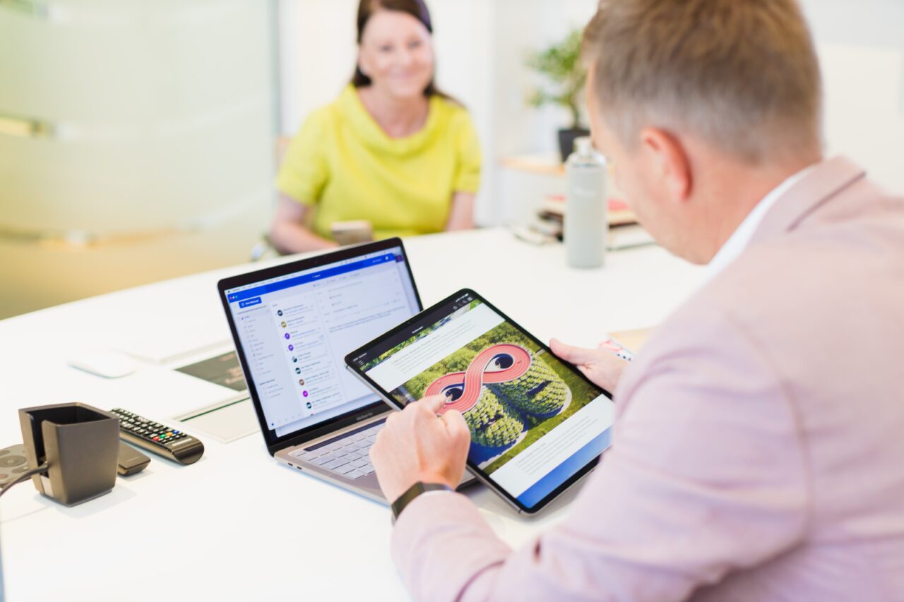 An over the shoulder shot of a male in a pink suit holding an electronic table over a white desk with a laptop and remote controls on top of it, There is a blurry female in the background