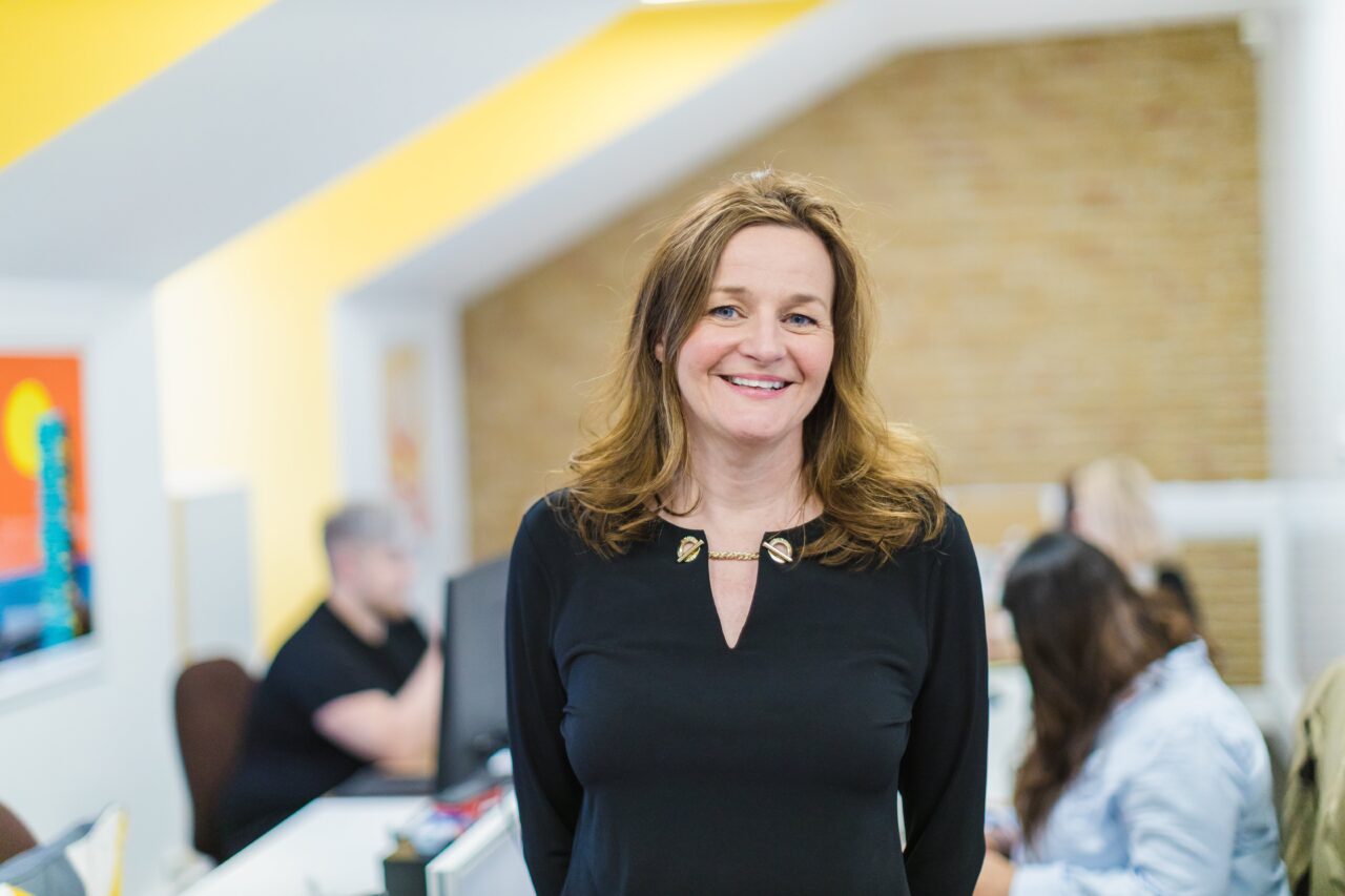 A female financial executive stands smiling in an office whilst three colleagues sit a desk behind her