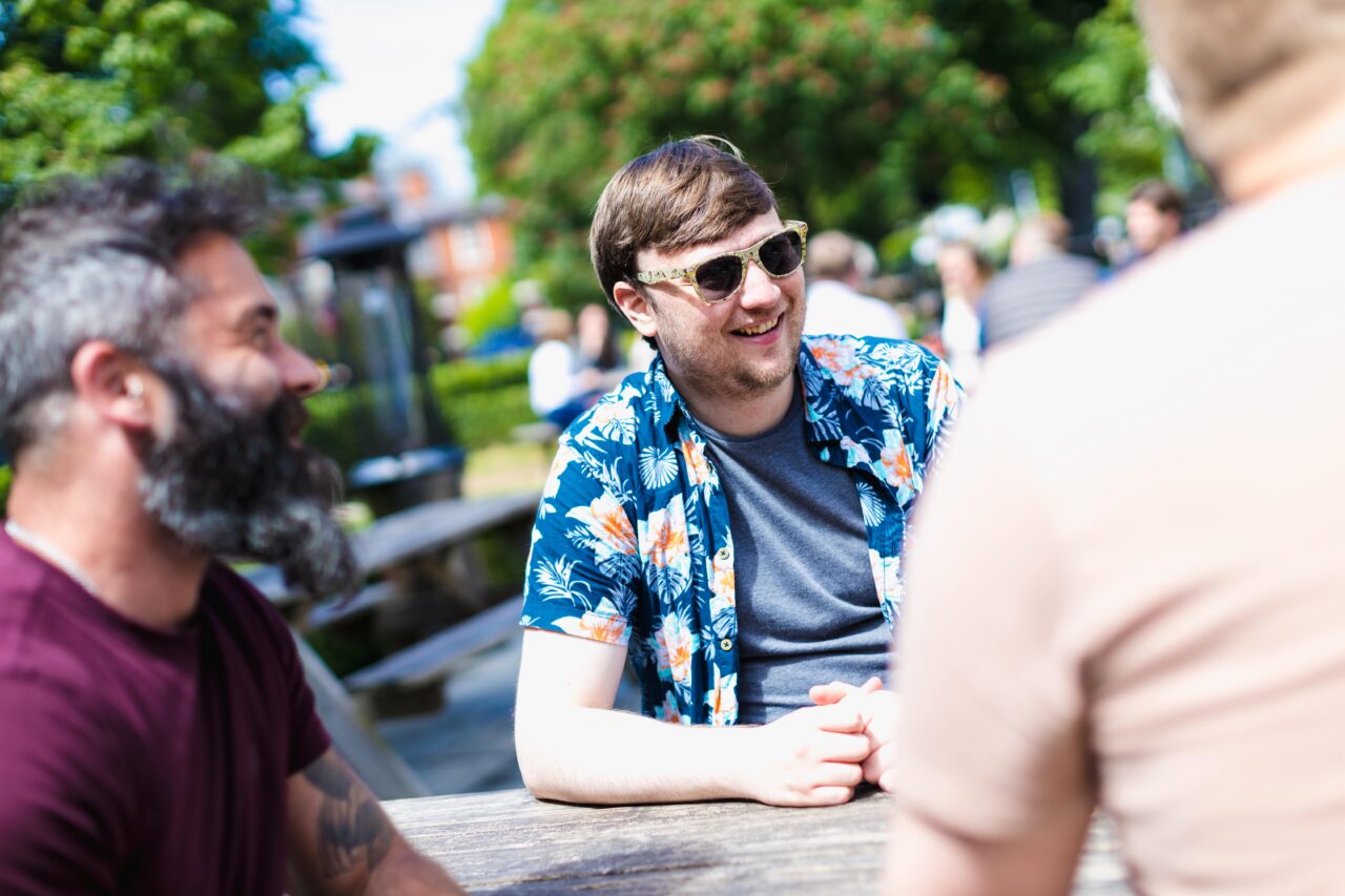 Three males sat in an outdoor area on a sunny day smiling, one is wearing sunglasses