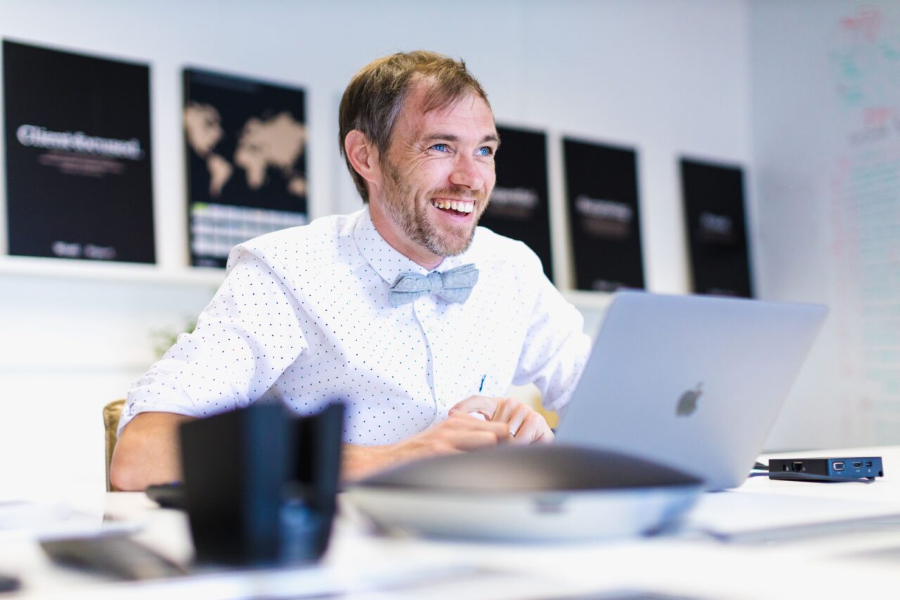 A male is sat at a desk in front of a laptop looking upwards and laughing