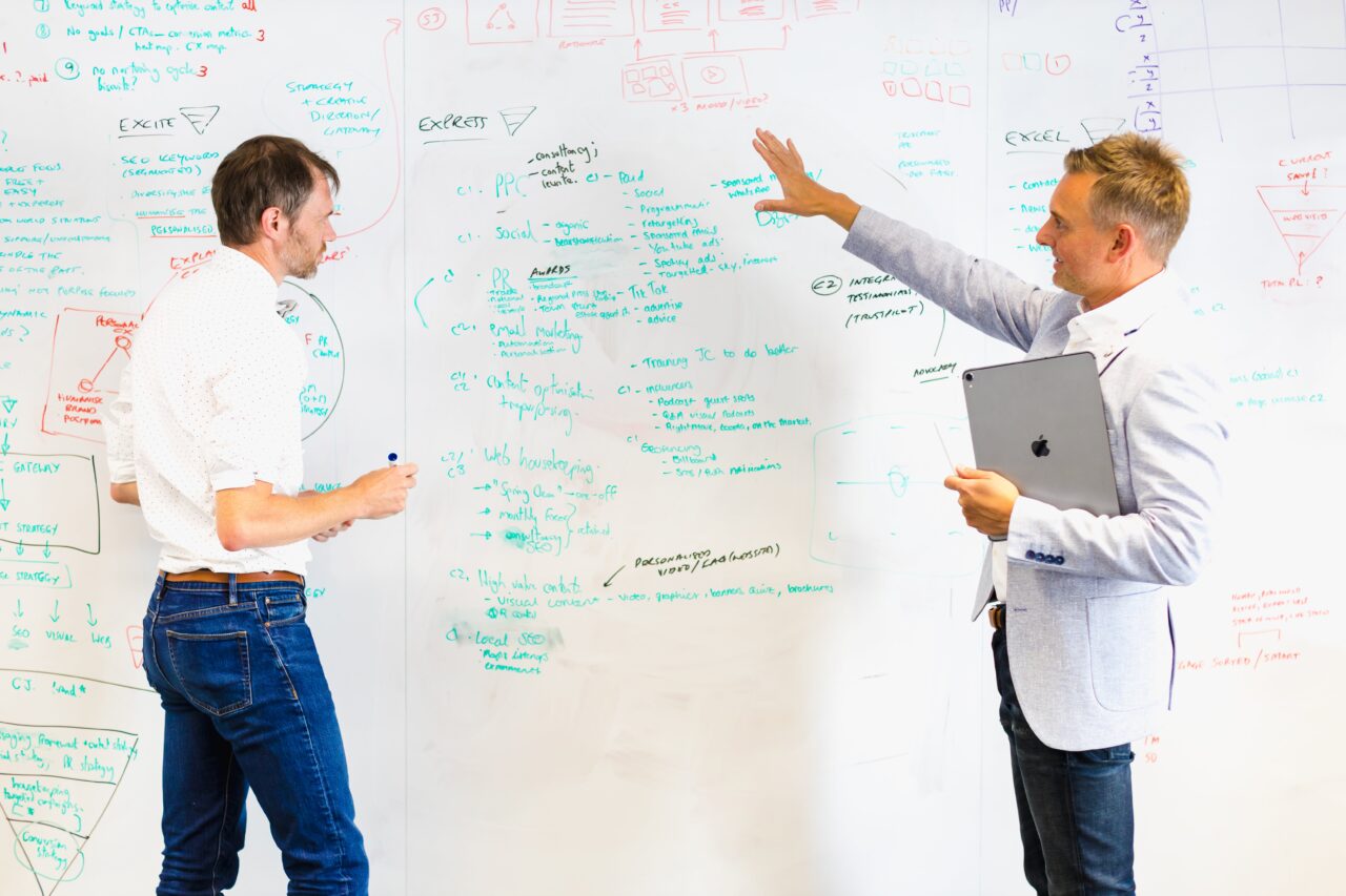 Two males stand in front of a whiteboard discussing the text written all over, one male is holding a pen and the other is holding an electronic tablet