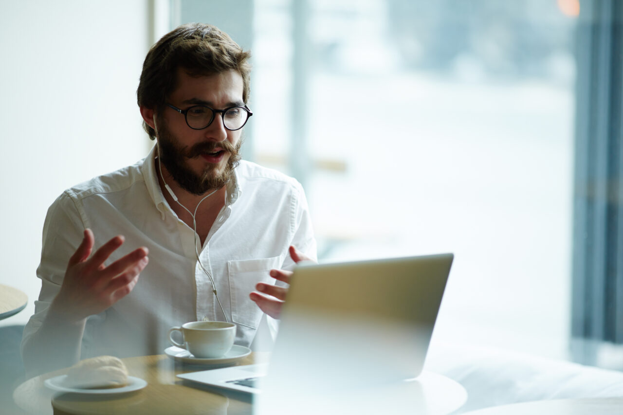 A mid height shot of a beared male sat at a desk wearing headphones, in front of a laptop and a coffee cup