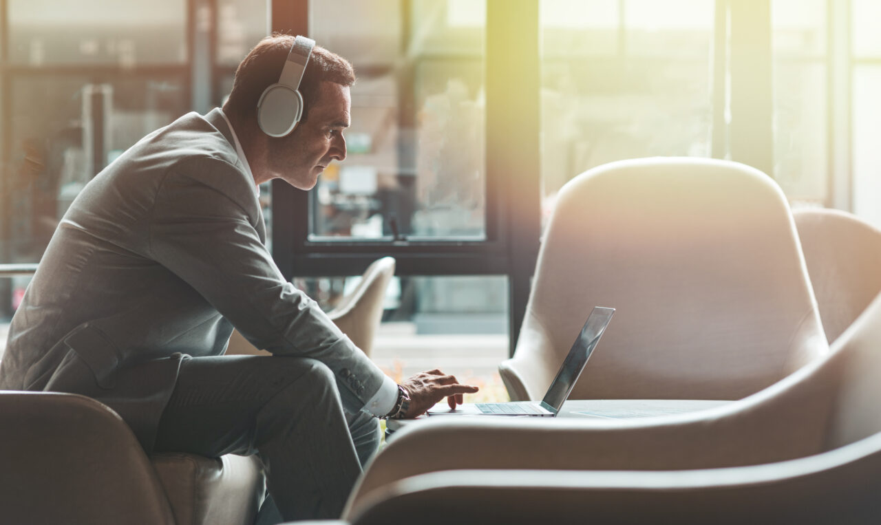 A male is sat in an armchair wearing headphones leaning over a laptop which is sat on a coffee table in front of him, sunshine is coming through the window in the background