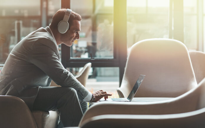 A male is sat in an armchair wearing headphones leaning over a laptop which is sat on a coffee table in front of him, sunshine is coming through the window in the background