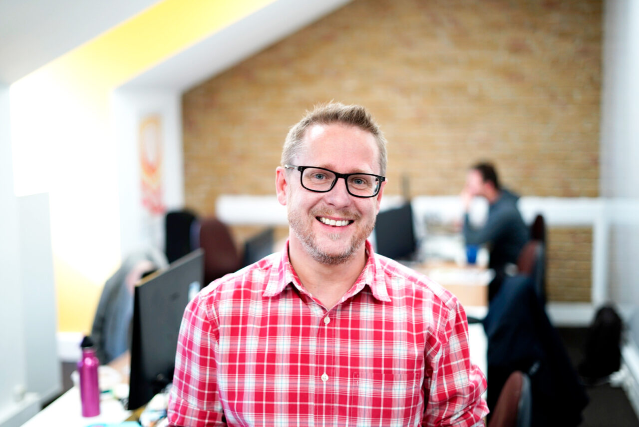 Matt Lakey, head of digital transformation and performance, standing in an office with a colleague sat at a desk in the background