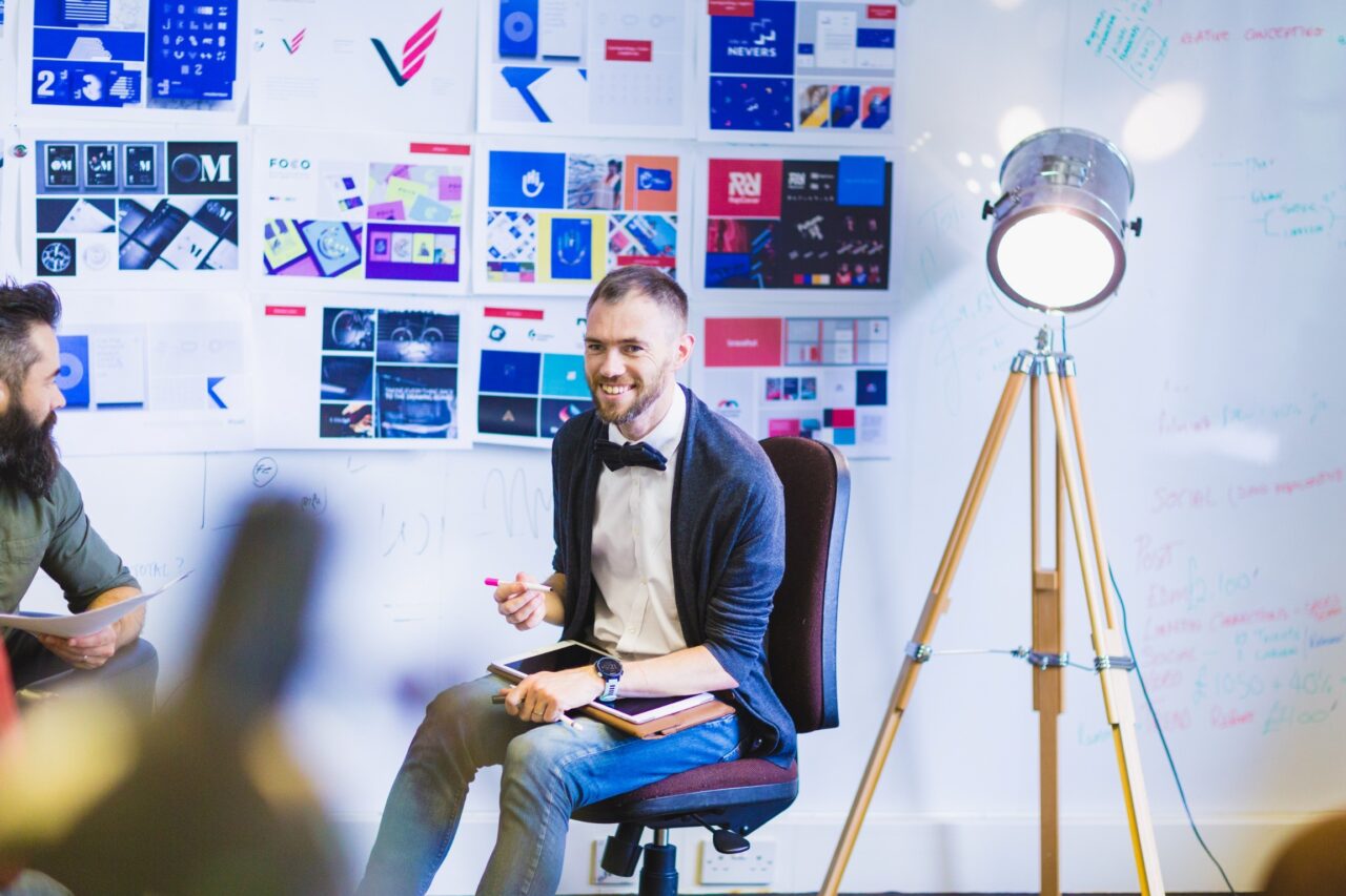 Two males are sat down in front a white board featuring graphic design work pasted all over it