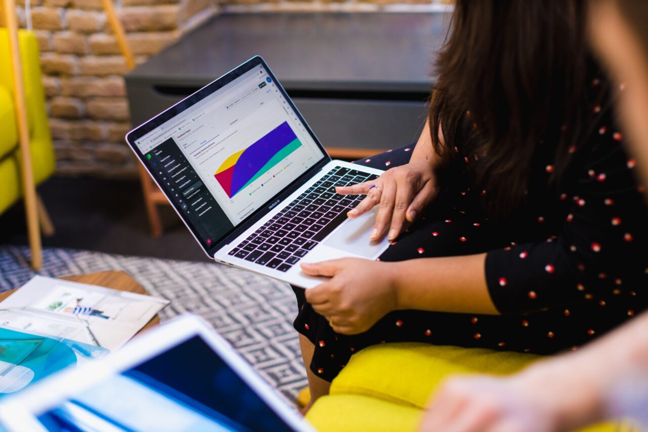 An over the shoulder shot of a female and male sat on a yellow couch, the female is holding a laptop with a presentation visible on screen
