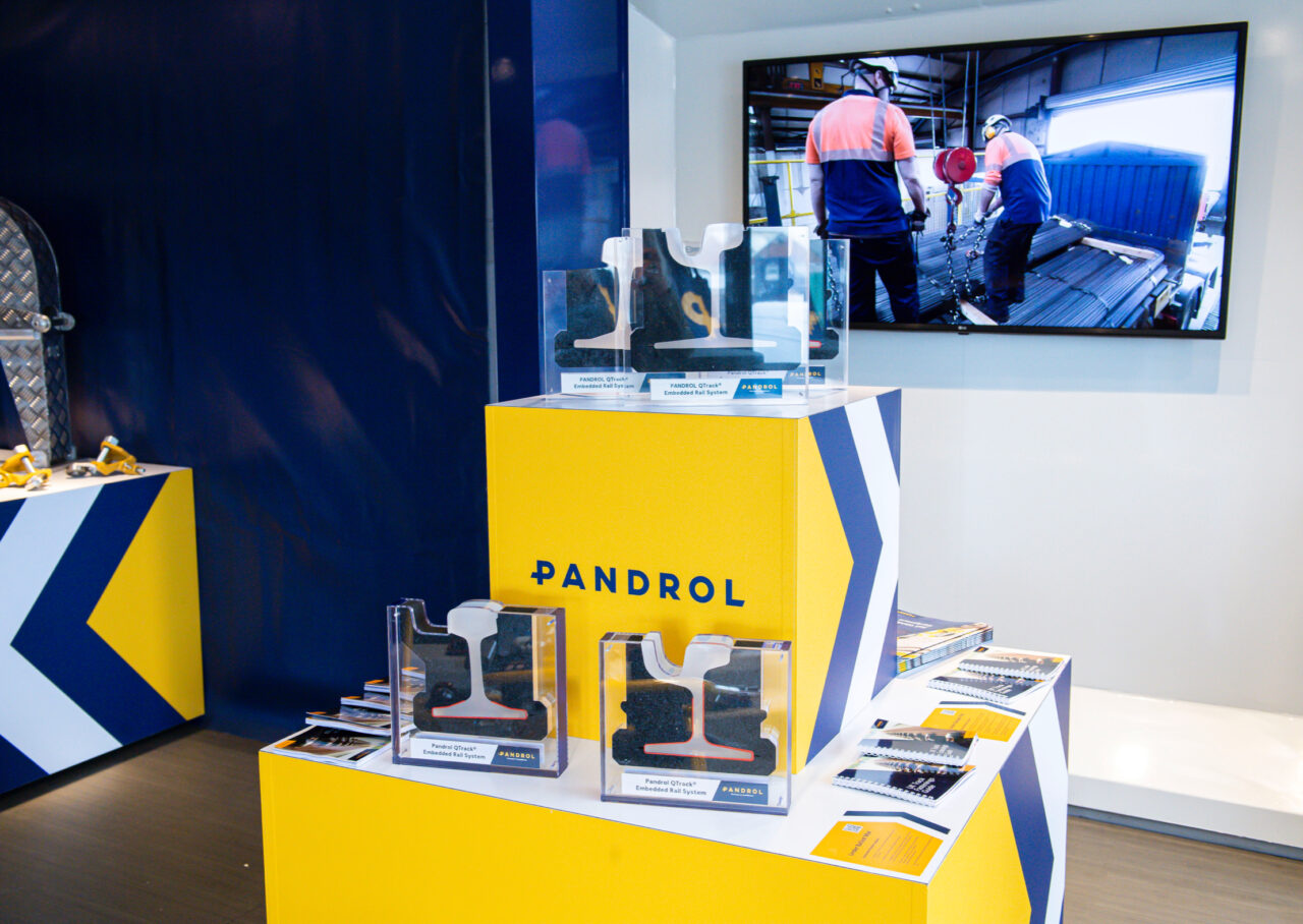 A Pandrol branded table with documentation on top, with a wall mounted tv behind showing warehouse workers at work
