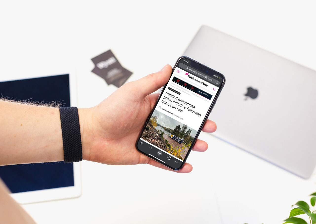 A hand holding a mobile phone showing a news article about Pandrol's green initiative, whilst a laptop sits on a desk in the background