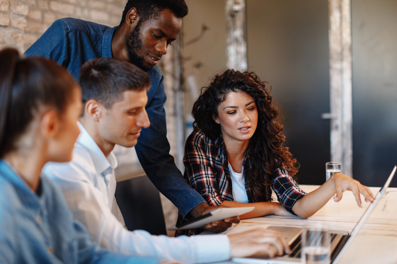 A mid height shot of two males and two females all looking at a laptop sitting on a brown desk