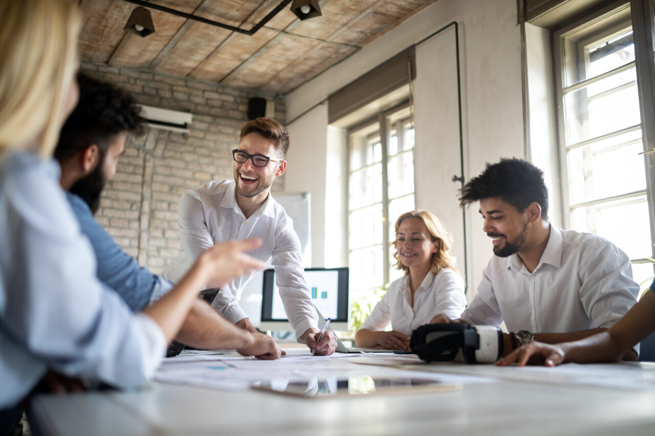 A mid height shot of a group of male and female office workers, sat at a white desk covered in pieces of paper, all people are smiling