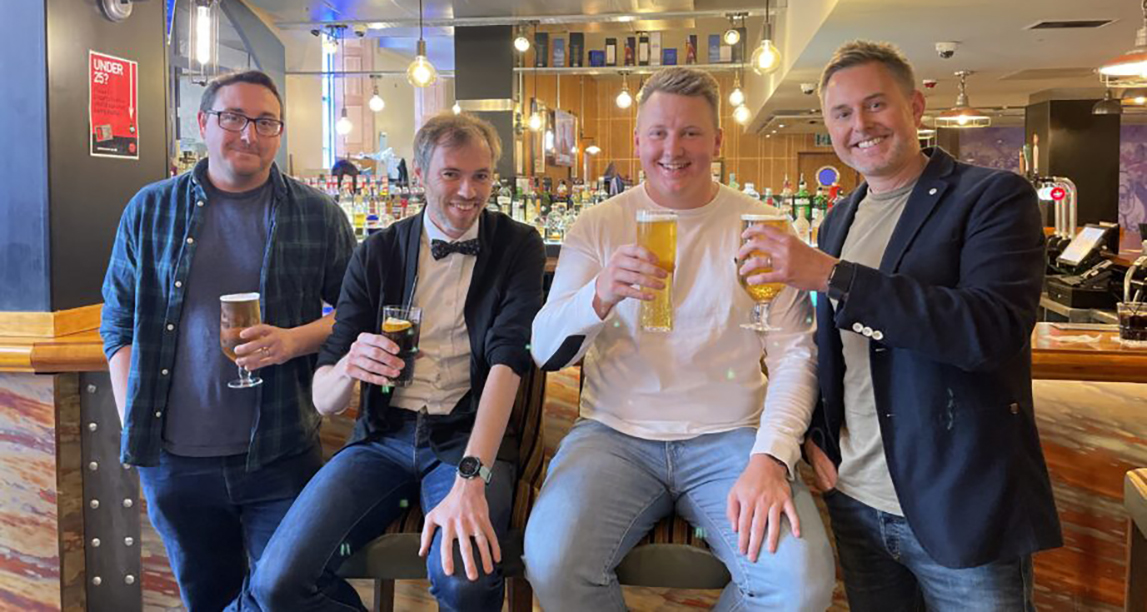 two men sitting and two men standing and smiling in a bar, all holding drinks