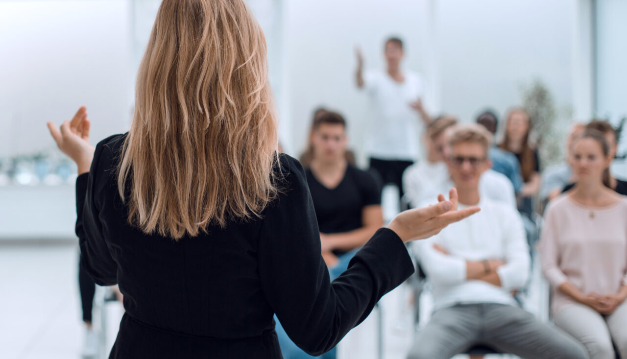 A mid-height, over the shoulder shot of a female delivering a speech to a group of people who are blurred in the background