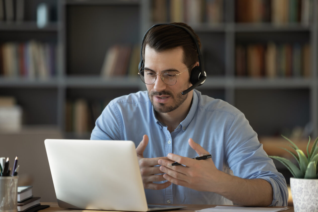 A zoomed in shot of a man wearing a headset and a blue shirt, sat behind a desk with a laptop, plant and pot of pens