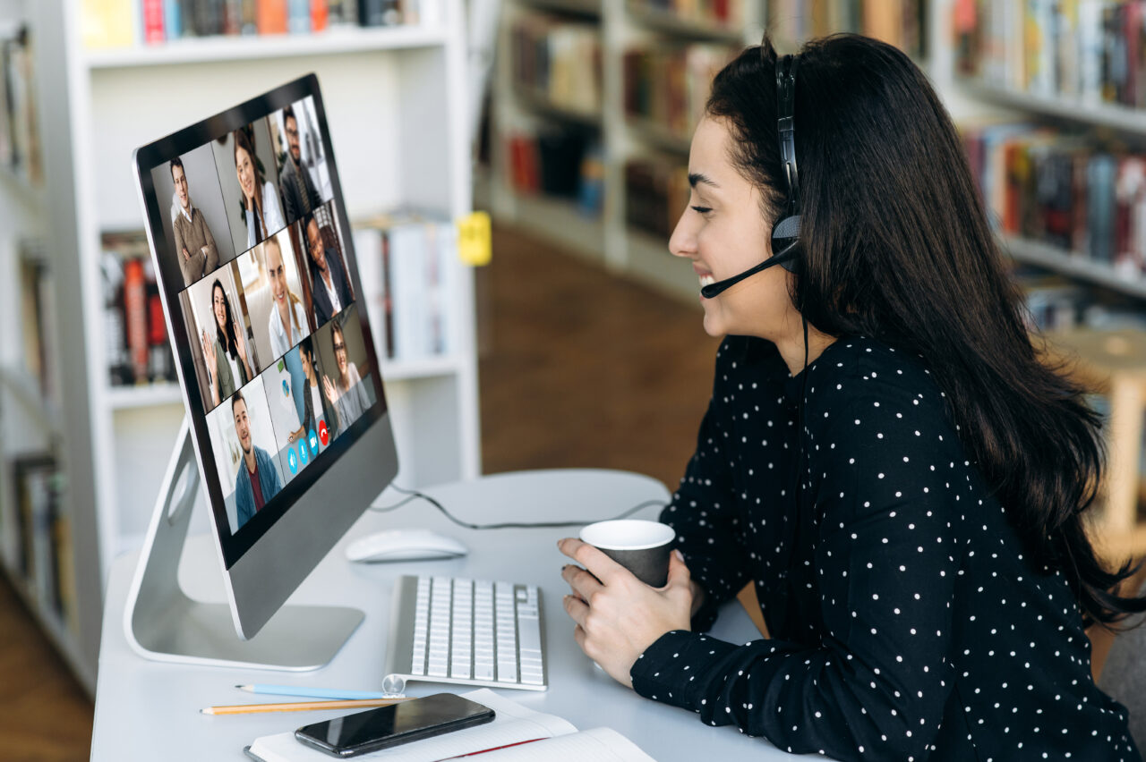 A female sitting at a desk wearing a headset and holding a cup whilst smiling at a computer screen