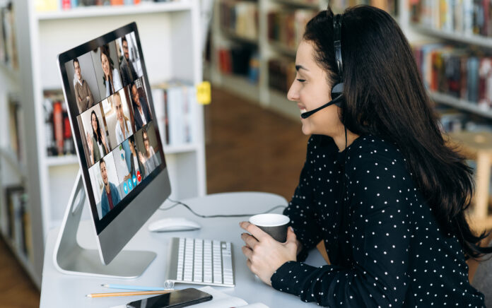 A female sitting at a desk wearing a headset and holding a cup whilst smiling at a computer screen