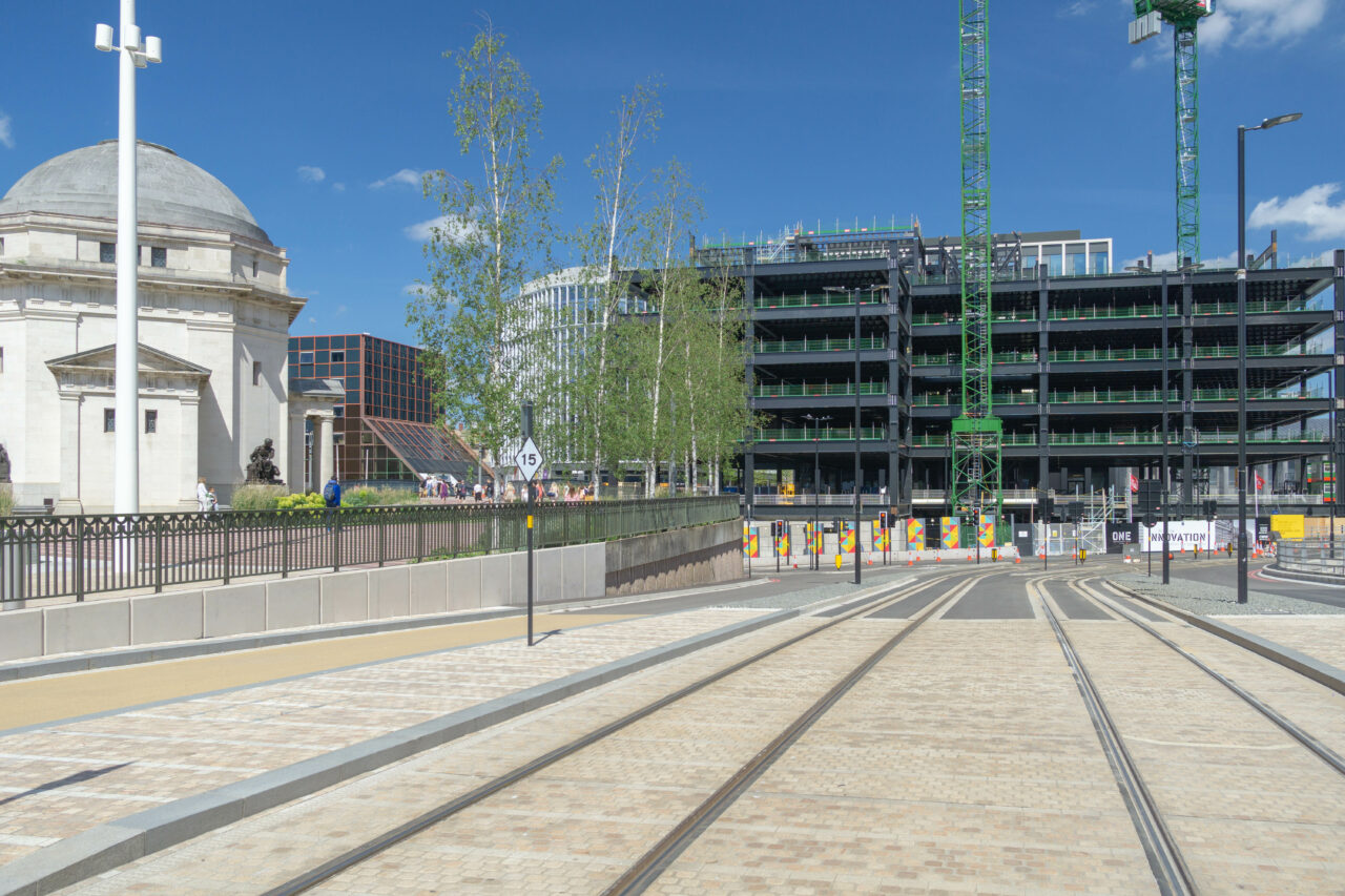 A wide day time shot of tramlines with construction of a building in the background and a monument to the left of the tramlines