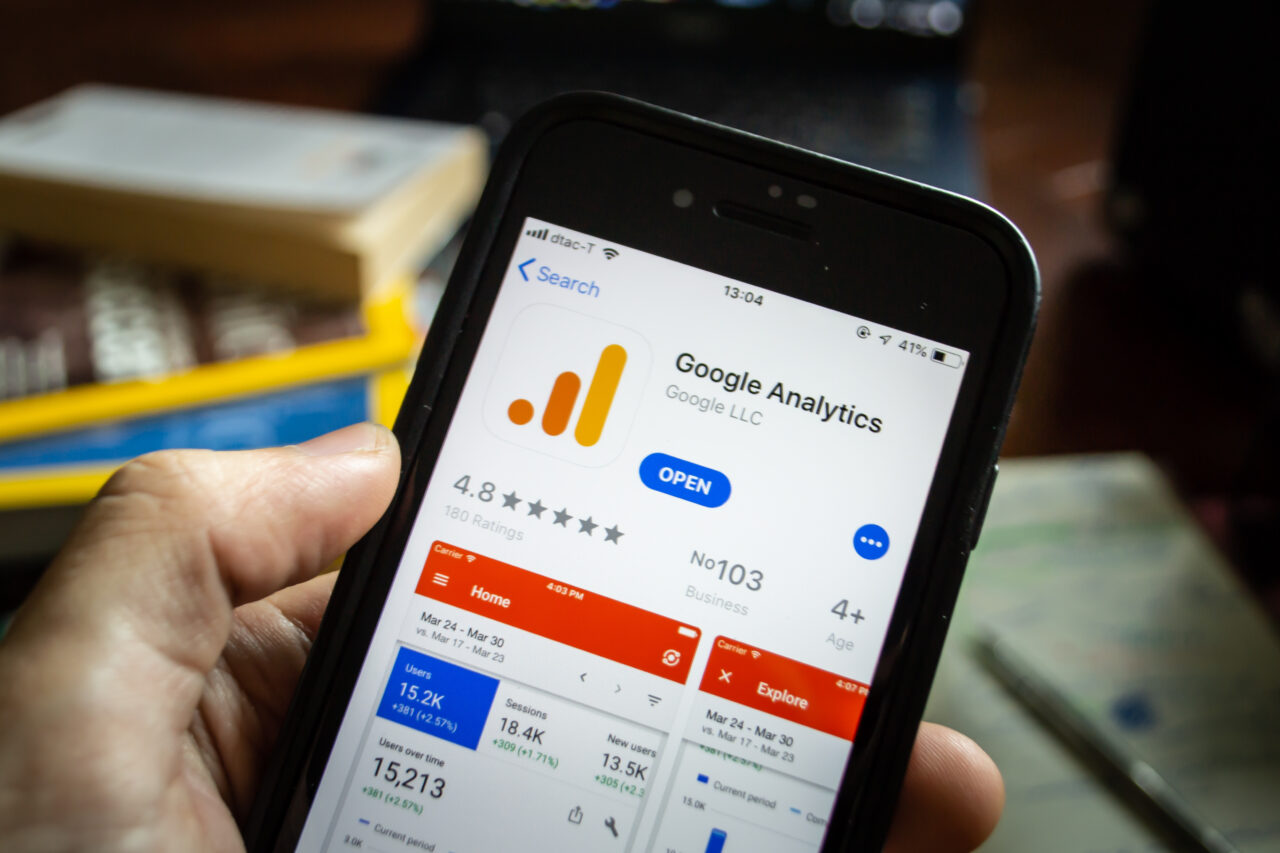 A close up shot of a mobile phone being held by a persons hand, with the google analytics app store listing on screen. There are blurry books and a desk in the background