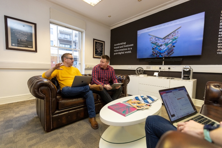 Two male office workers sat on a brown leather sofa behind a white coffee table. Both males are holding a laptop and an electronic tablet