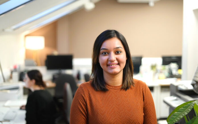 Sahar Afzal, Brand & Marketing Strategist, stood in an office smiling whilst a colleague sits at a desk behind her.