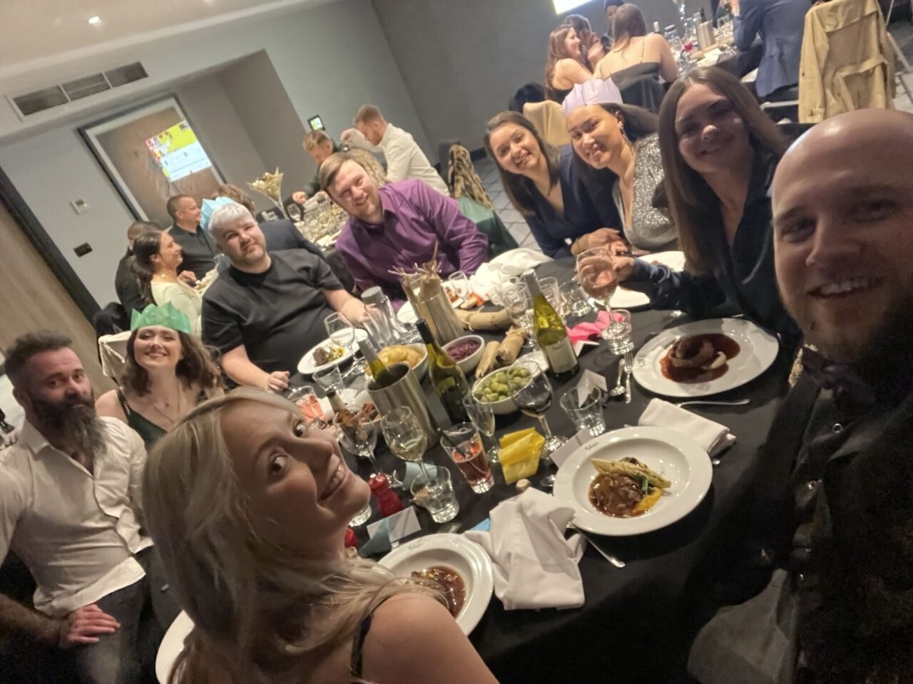 A Christmas party, with office workers sat at a dinner table with plates of food and bottles of wine