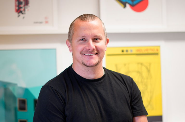 Toby Aiken, Strategic Account Director, is stood smiling in front of creative design work