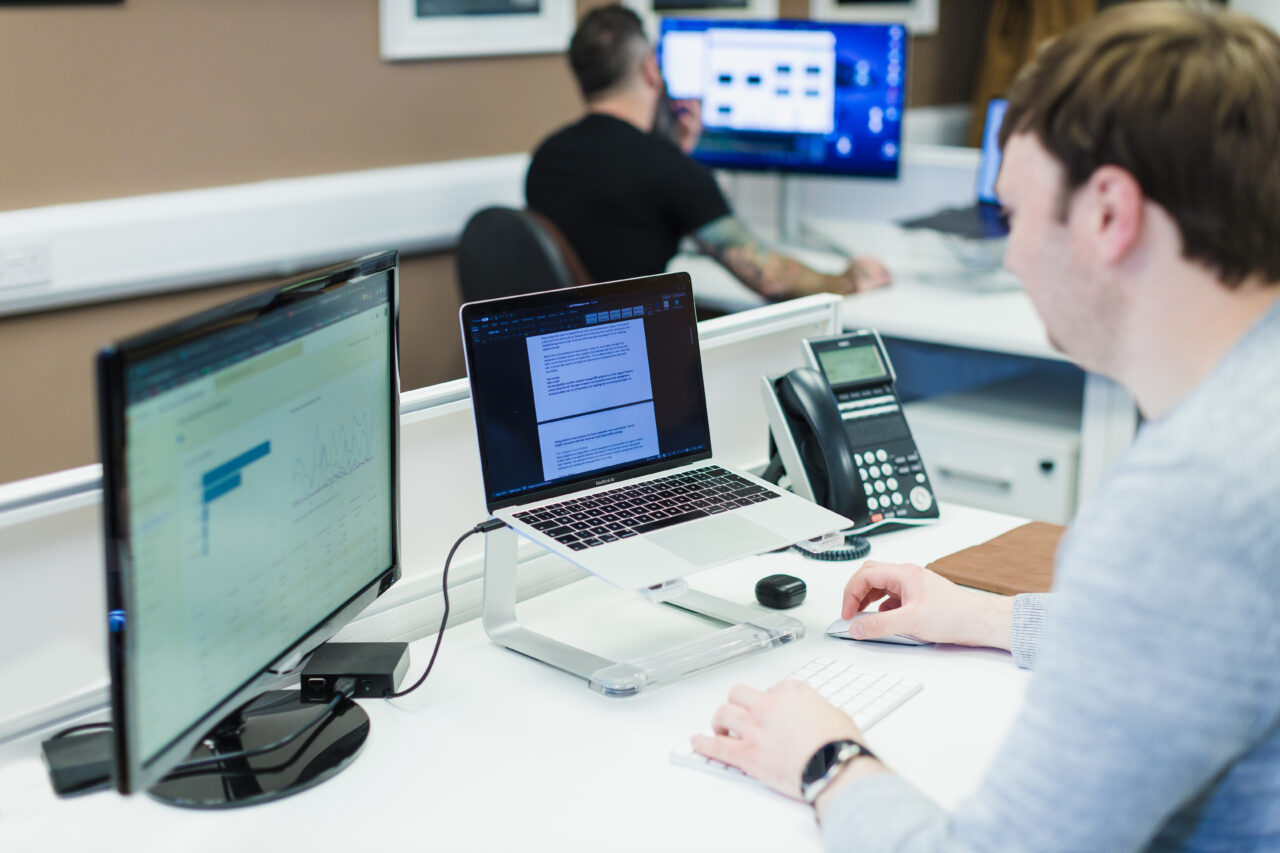 A shot of a mac book and secondary monitor and a male working at his desk.