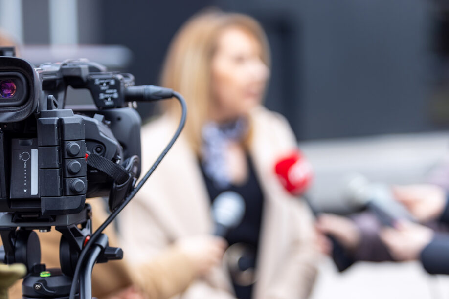 A video camera is actively recording as a journalist reports on a current news event